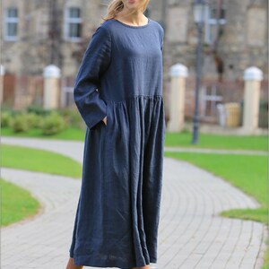 May include: A woman wearing a long, dark blue linen dress with a gathered waist and long sleeves. She is wearing brown lace-up boots and is standing on a brick pathway.
