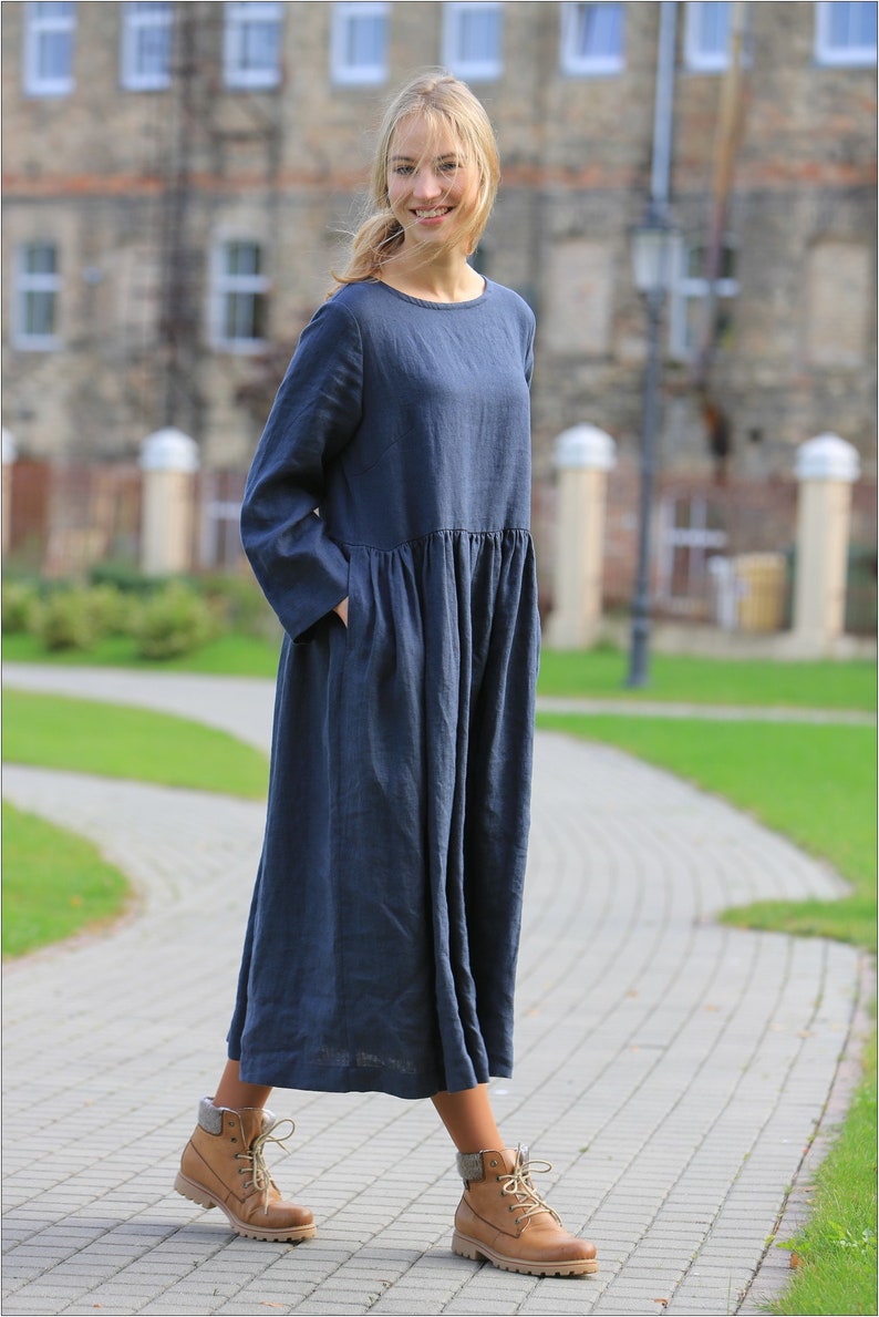May include: A woman wearing a long, dark blue linen dress with a gathered waist and long sleeves. She is wearing brown lace-up boots and is standing on a brick pathway.