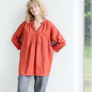 May include: A loose-fitting, terracotta-colored linen top with a V-neck and a gathered neckline. The top has long sleeves and a relaxed fit.