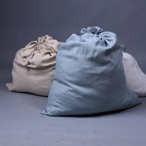 Pode incluir: Três sacos de tecido com cordão de ajuste em diferentes cores: bege, azul claro e branco. Os sacos são feitos de um material macio e natural e são perfeitos para guardar ou transportar objetos.