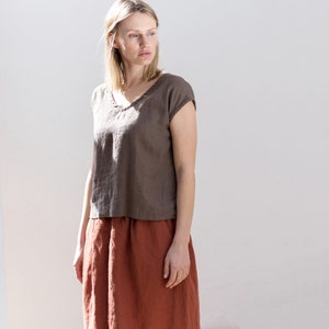 May include: A woman wearing a brown linen top with a v-neck and short sleeves, paired with a rust-colored linen skirt.