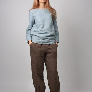May include: A woman wearing a light blue linen top and brown linen pants with cuffed hems. She is wearing brown leather shoes with tan laces.