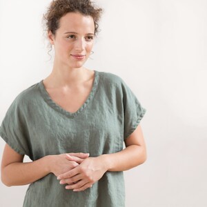 May include: A woman wearing a loose-fitting, sage green linen top with a v-neckline. The top has short sleeves and a relaxed fit.