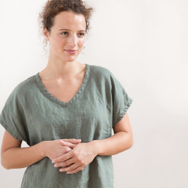 May include: A woman wearing a loose-fitting, sage green linen top with a v-neckline. The top has short sleeves and a relaxed fit.