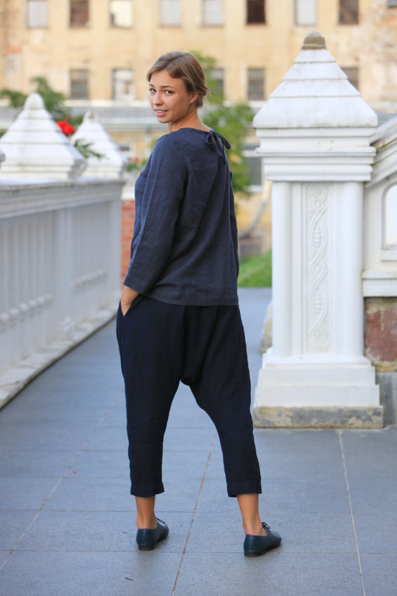 May include: A woman wearing a dark blue long-sleeved shirt and dark blue pants with a loose, baggy fit. She is standing on a paved surface in front of a white stone structure.