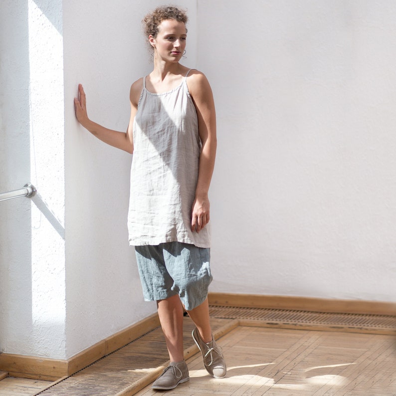 May include: A woman wearing a light beige linen tank top and blue linen shorts. She is standing in a room with white walls and wooden floors. She is looking to the right of the frame.