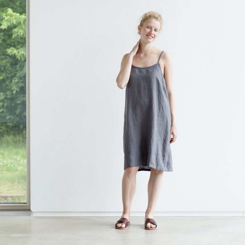 May include: A woman wearing a gray linen dress with thin straps. She is standing in front of a white wall and smiling at the camera.
