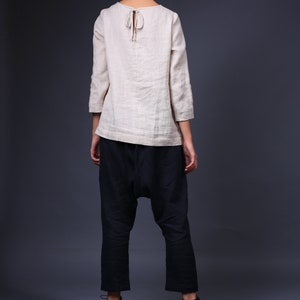 May include: A woman wearing a light beige linen top with a tie at the back and dark blue pants. She is standing on a wooden pallet.