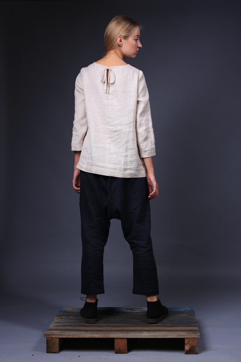 May include: A woman wearing a light beige linen top with a tie at the back and dark blue pants. She is standing on a wooden pallet.