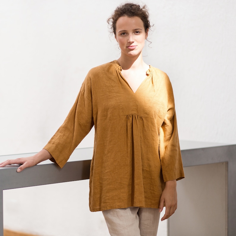 May include: A woman wearing a loose-fitting, mustard yellow linen top with a V-neck and 3/4 sleeves. She is standing in front of a white wall and is leaning on a gray countertop.