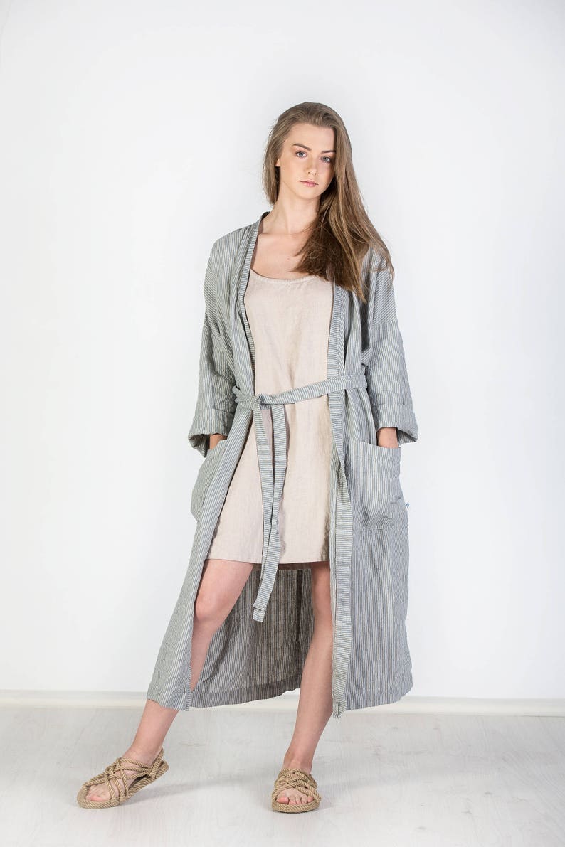 May include: A woman wearing a long, striped, gray linen robe with a matching belt. She is wearing a beige linen dress underneath the robe. She is standing on a white background and wearing sandals.