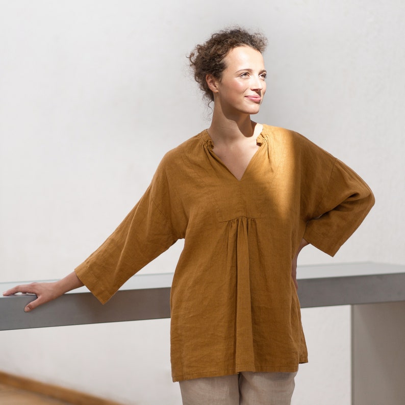 May include: A woman wearing a loose-fitting, mustard yellow linen top with a V-neck and 3/4 sleeves. The top has a gathered detail at the waist.