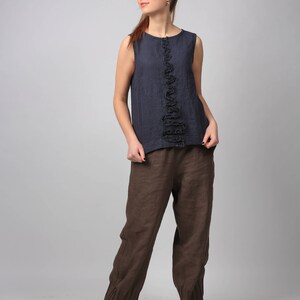 Puede incluir: Una mujer con un top azul oscuro sin mangas con un detalle de volantes en la parte delantera y pantalones de lino marrones. Lleva botas de cuero marrones.