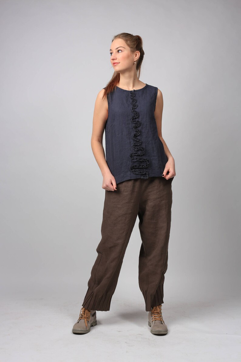 Puede incluir: Una mujer con un top azul oscuro sin mangas con un detalle de volantes en la parte delantera y pantalones de lino marrones. Lleva botas de cuero marrones.