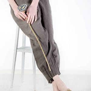 May include: A person wearing brown linen pants and beige rope sandals sits on a white stool. They are holding a dried lotus pod.
