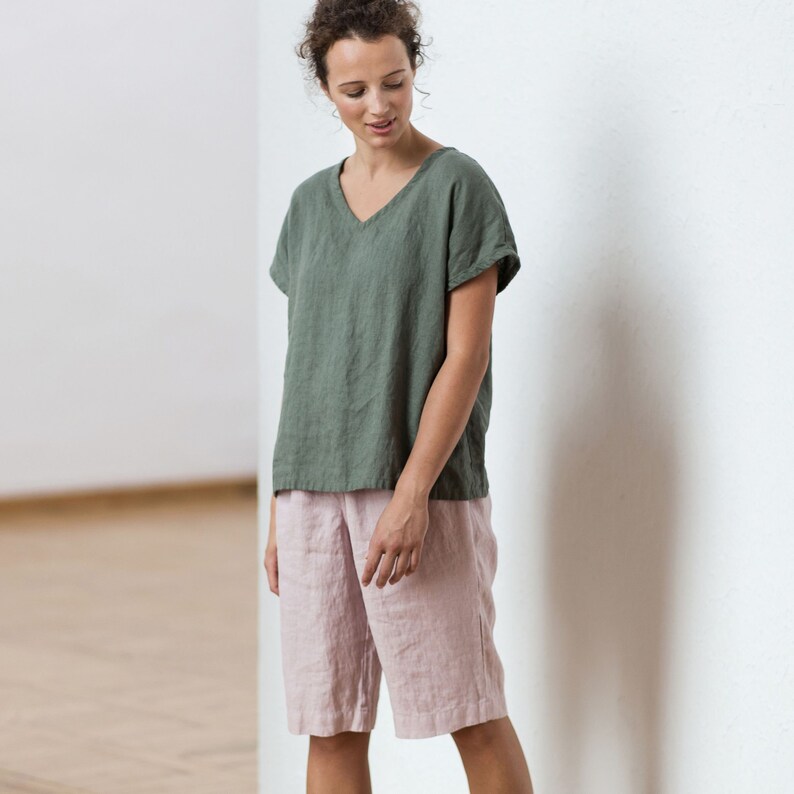 May include: A woman is wearing a green linen top with a v-neck and short sleeves. She is also wearing pink linen shorts.