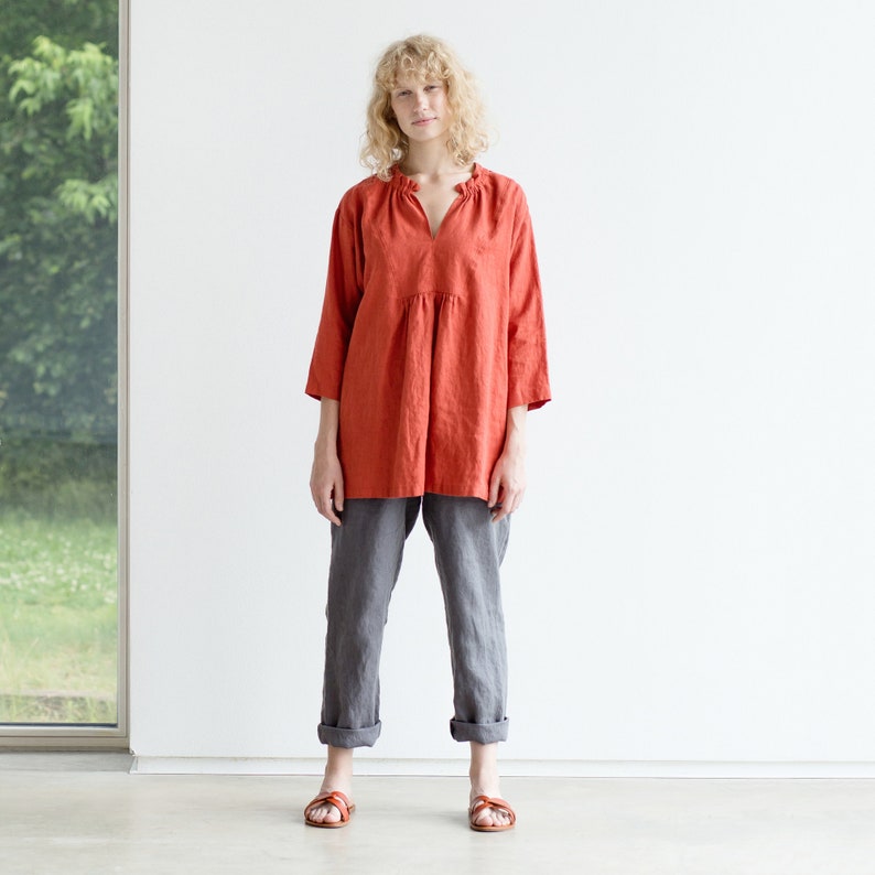 May include: A woman wearing a loose-fitting, rust-colored linen top with a ruffled neckline and grey linen pants. She is standing on a white background.