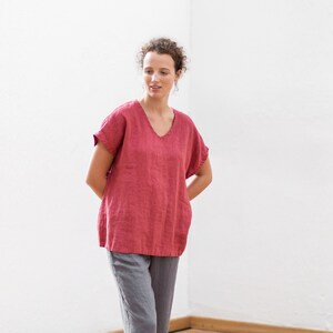 May include: A woman wearing a loose-fitting, red linen top with a v-neck and short sleeves. She is wearing gray pants.