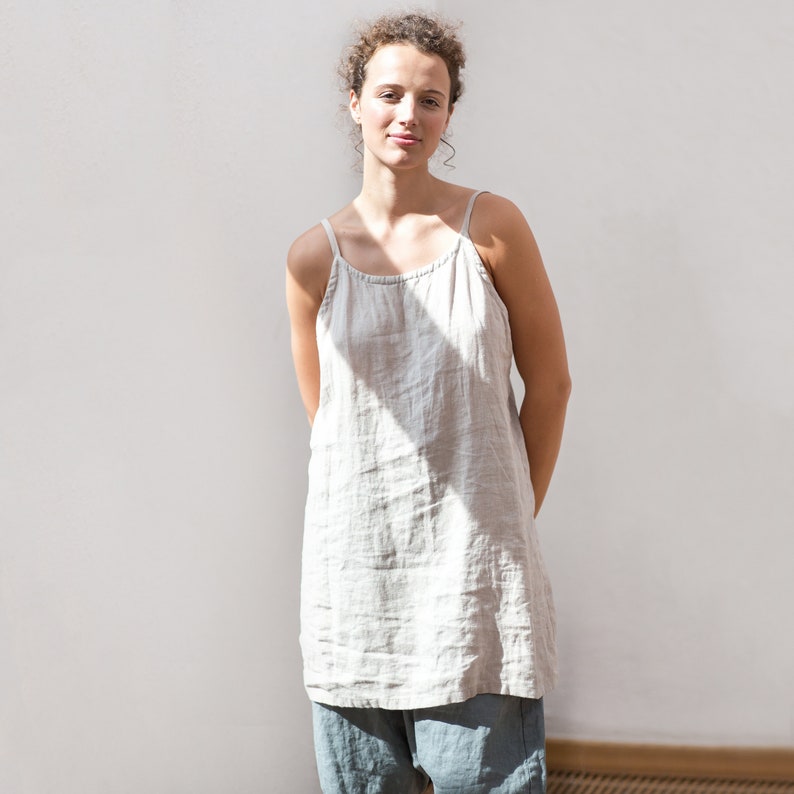 May include: A woman wearing a light beige linen tank top with a relaxed fit. The top has thin straps and a scoop neck. The woman is standing in front of a white wall.