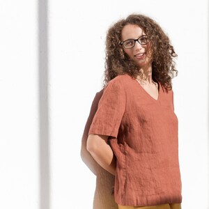 May include: A woman with curly brown hair wearing glasses and a rust-colored linen top.