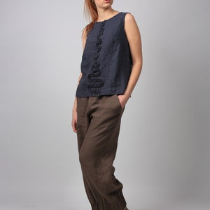 May include: A woman wearing a dark blue sleeveless top with a black ruffle detail and brown linen pants. She is wearing gray canvas shoes with brown laces.