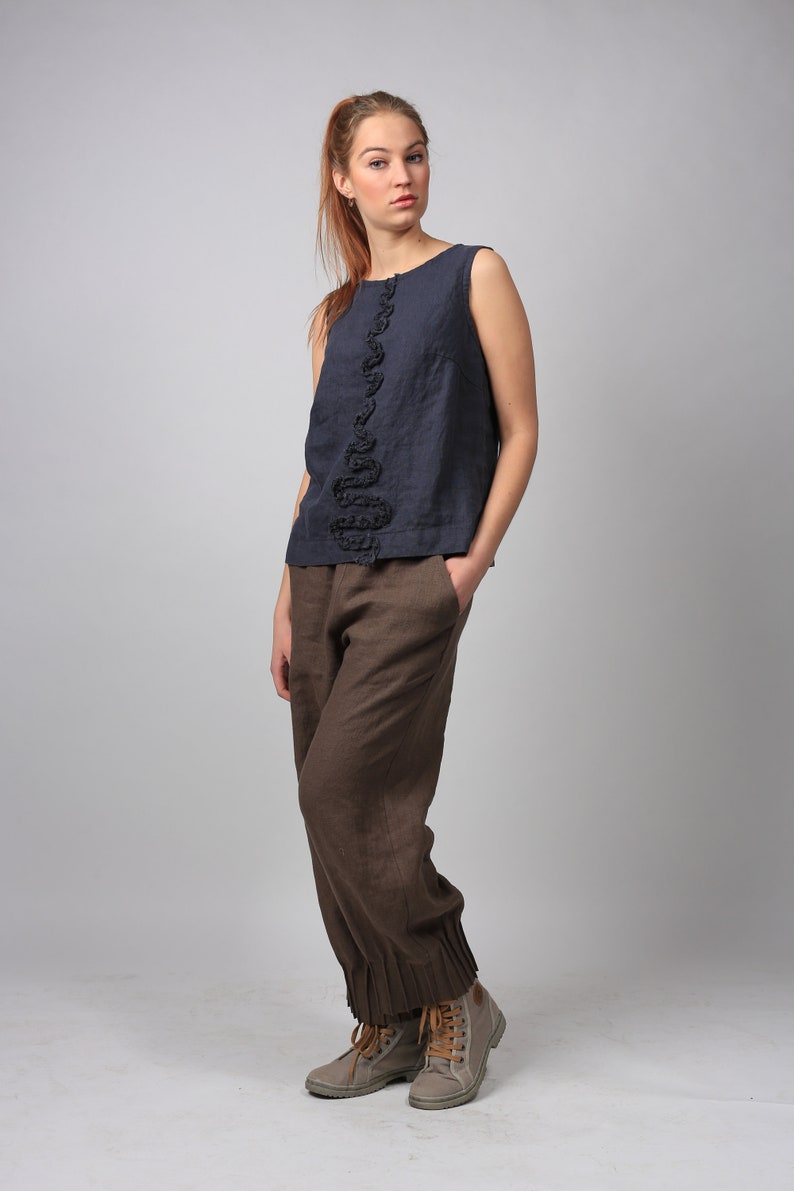 May include: A woman wearing a dark blue sleeveless top with a black ruffle detail and brown linen pants. She is wearing gray canvas shoes with brown laces.