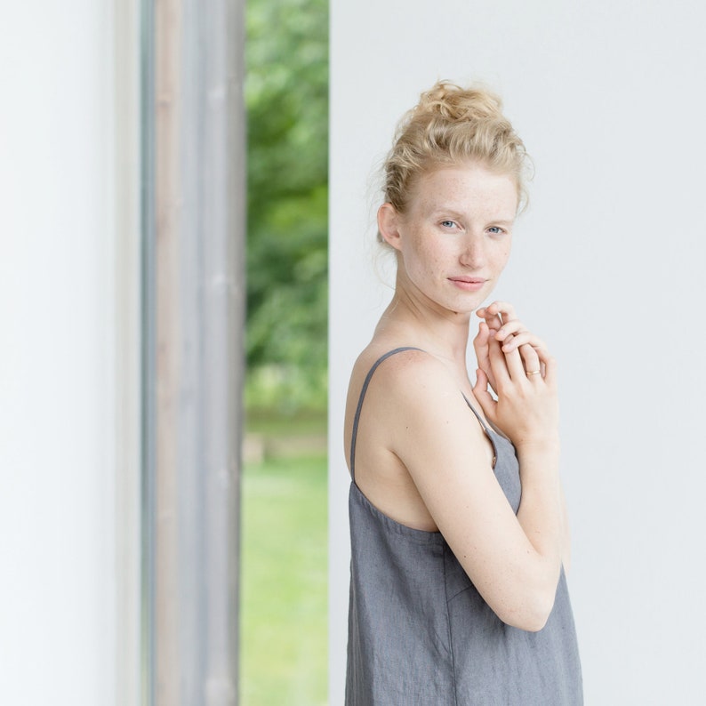 May include: A woman with blonde hair pulled back in a bun is wearing a gray sleeveless linen dress. She is standing in front of a window with a white wall behind her. She is looking at the camera with her hands clasped in front of her.