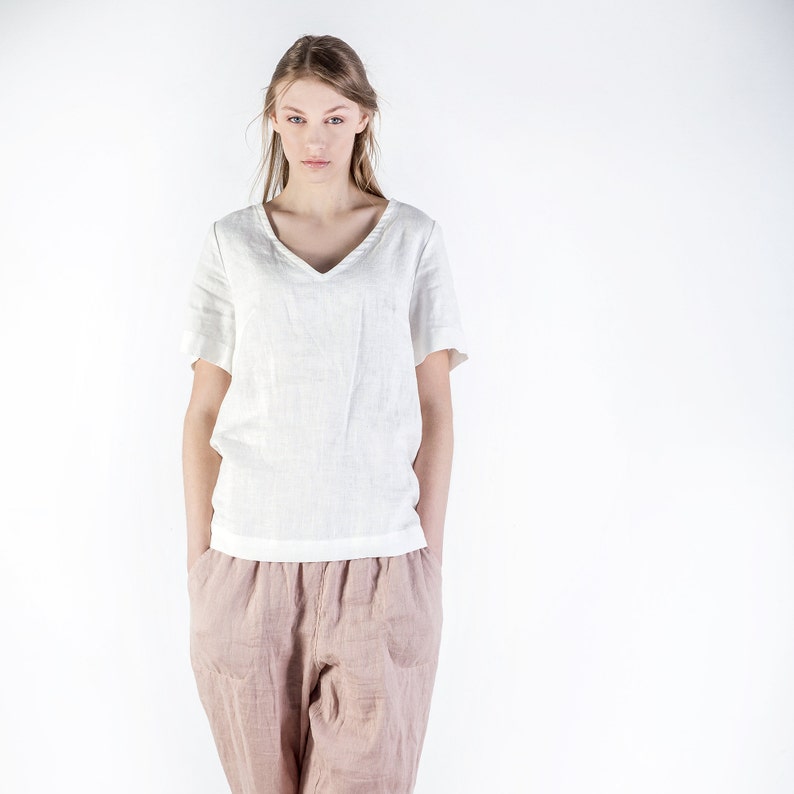 May include: A woman wearing a white linen top with a v-neck and a pair of light pink linen pants.