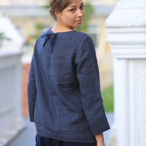May include: A woman wearing a dark blue linen top with a tie at the back of the neck. The top has long sleeves and a relaxed fit. She is wearing dark blue pants.