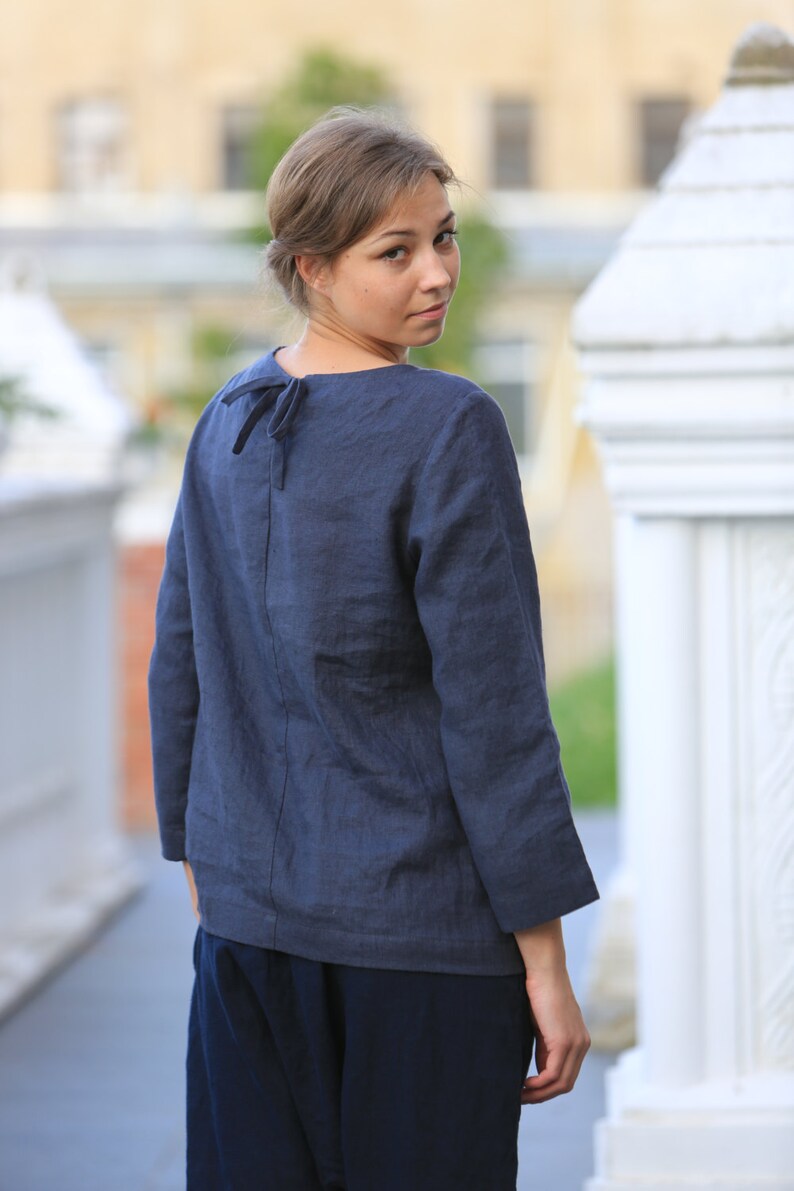 May include: A woman wearing a dark blue linen top with a tie at the back of the neck. The top has long sleeves and a relaxed fit. She is wearing dark blue pants.