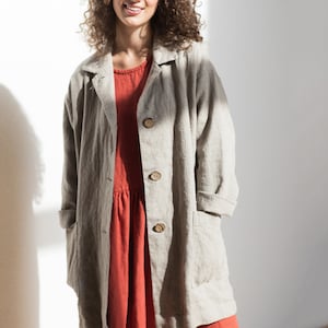May include: A woman wearing a light gray linen jacket over a red linen dress. The jacket has a collar, long sleeves, and four buttons down the front. The dress has a gathered skirt and a simple neckline.