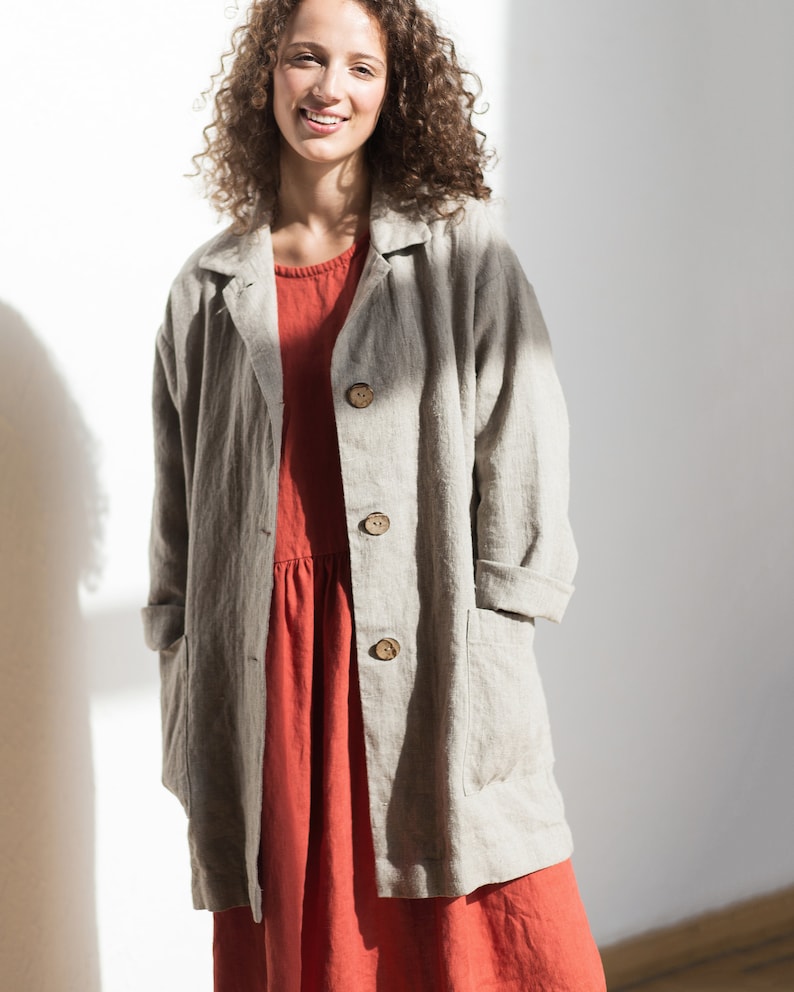 May include: A woman wearing a light gray linen jacket over a red linen dress. The jacket has a collar, long sleeves, and four buttons down the front. The dress has a gathered skirt and a simple neckline.