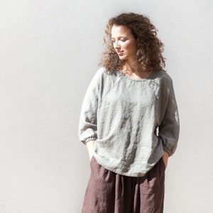 May include: A woman wearing a light gray linen top with a round neckline and three-quarter sleeves. She is also wearing a brown skirt.