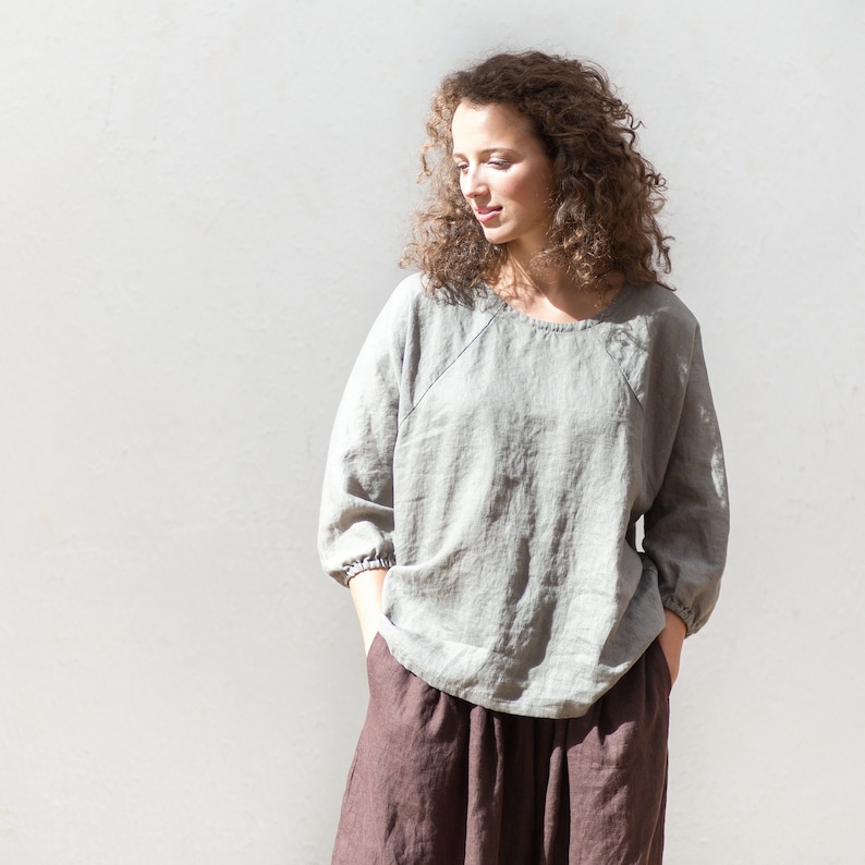 May include: A woman wearing a light gray linen top with a round neckline and three-quarter sleeves. She is also wearing a brown skirt.