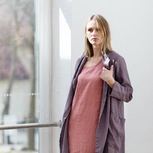 May include: A woman wearing a long, dusty rose-coloured linen dress with a matching linen cardigan in a darker shade of purple. The cardigan is open and the woman is holding the collar with her right hand.