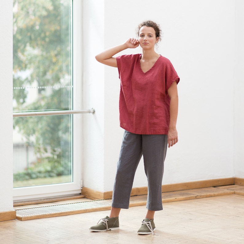 May include: A woman wearing a loose-fitting, red linen top with a V-neck and short sleeves. She is also wearing gray linen pants and olive green ankle boots.