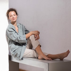 May include: A woman wearing a light blue linen shirt and beige linen pants sits on a bench with her legs crossed. She is looking to the side with a relaxed expression.
