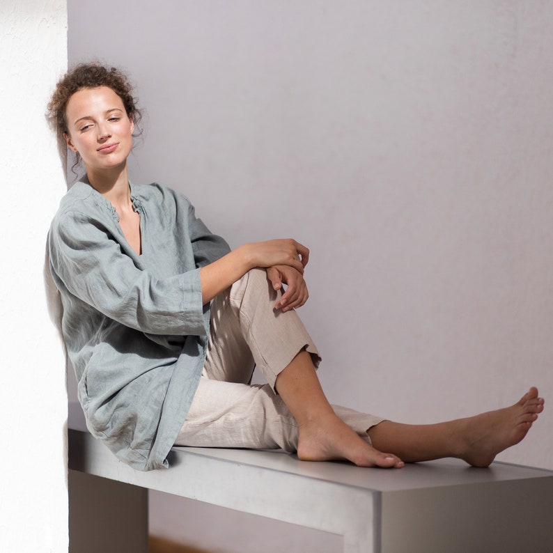 May include: A woman wearing a light blue linen shirt and beige linen pants sits on a bench with her legs crossed. She is looking to the side with a relaxed expression.