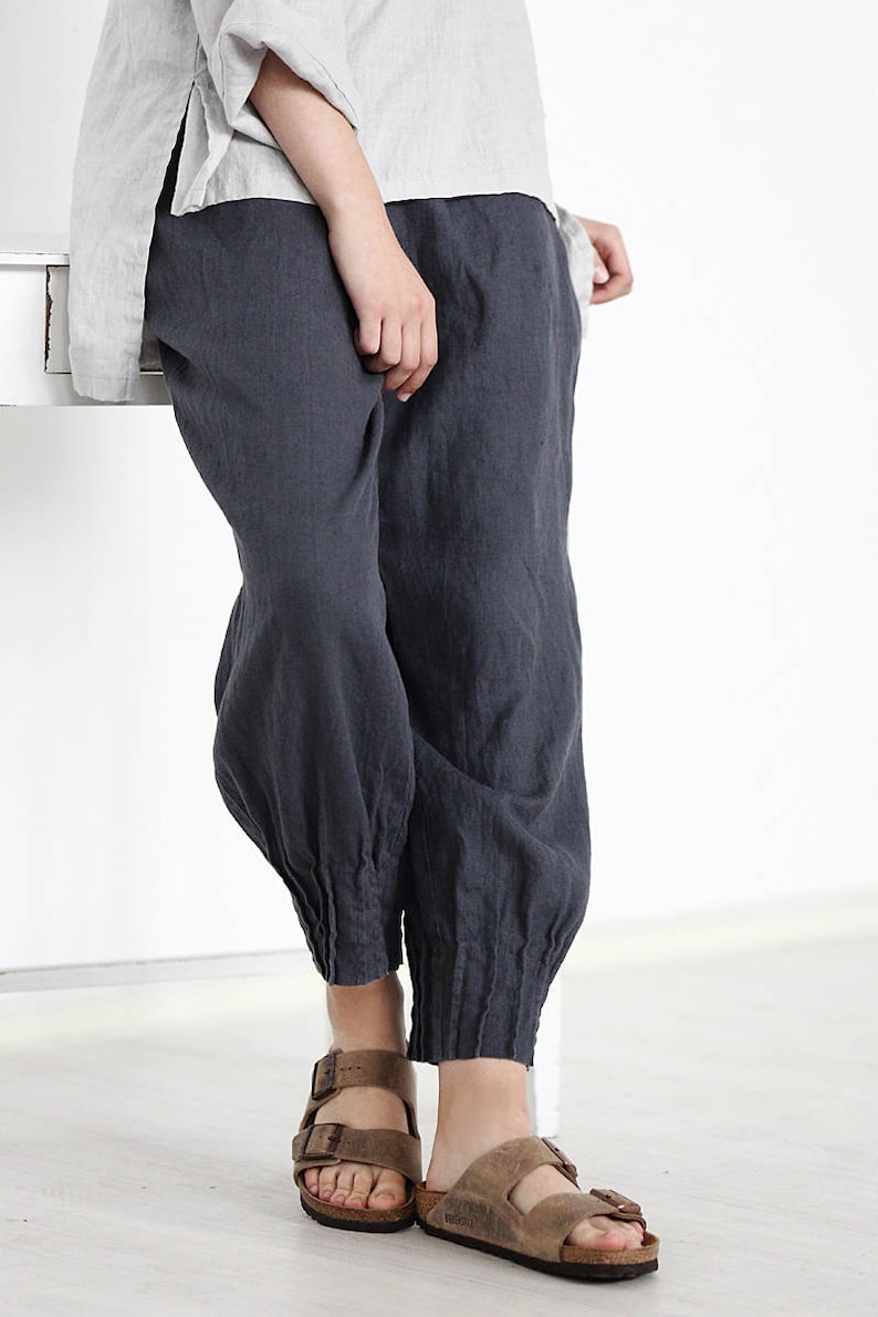 Puede incluir: Pantal&oacute;n de lino gris con un pu&ntilde;o el&aacute;stico fruncido en el tobillo. El pantal&oacute;n se lleva con sandalias de cuero marr&oacute;n.