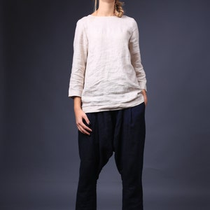 May include: A woman wearing a white linen top and dark blue, loose-fitting pants. She is standing on a wooden platform and looking directly at the camera.