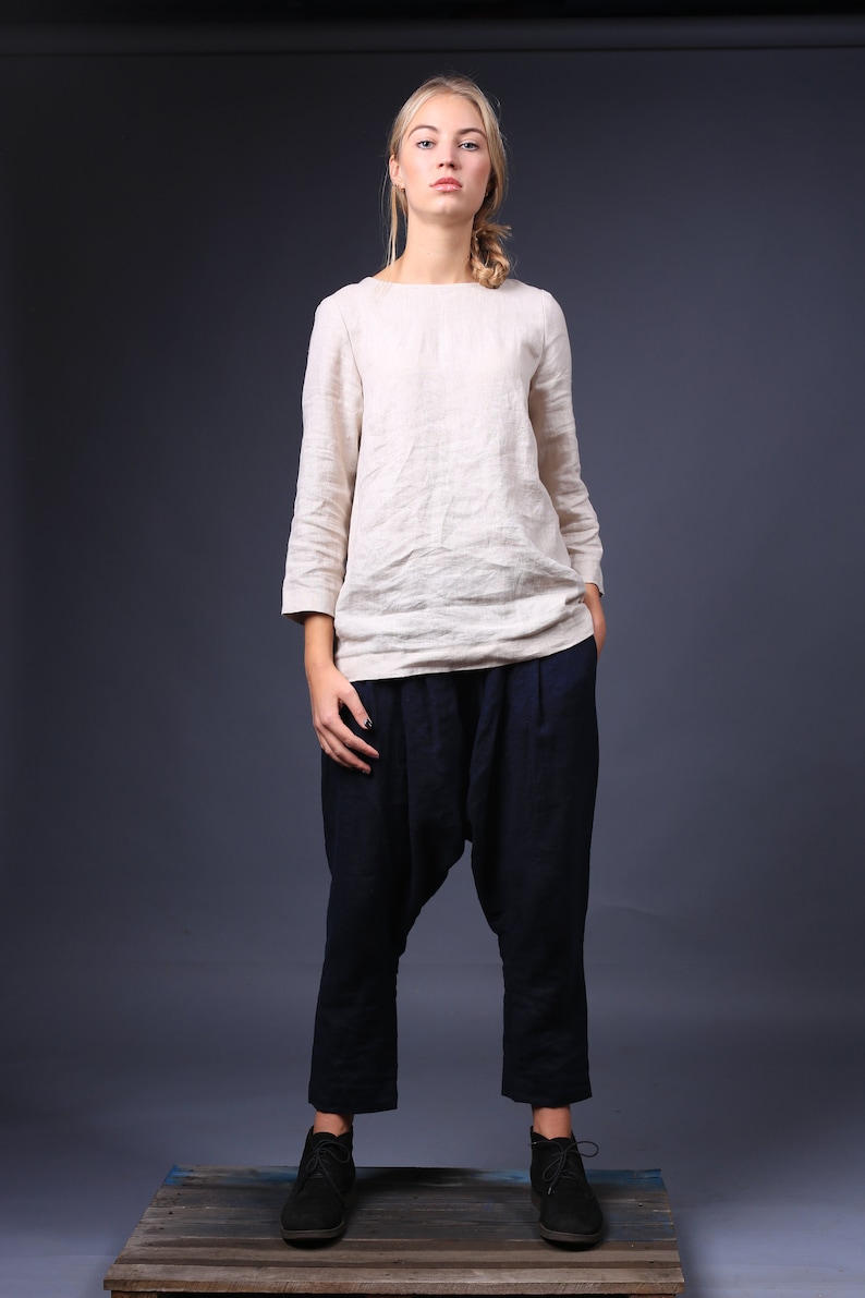 May include: A woman wearing a white linen top and dark blue, loose-fitting pants. She is standing on a wooden platform and looking directly at the camera.