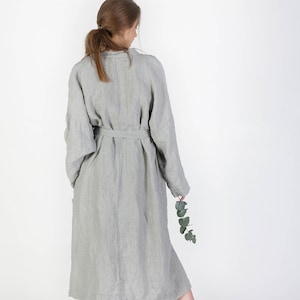 May include: A long, striped linen robe with a sash tie. The robe is a light gray color with thin, dark gray stripes. The robe is worn by a person standing with their back to the camera.
