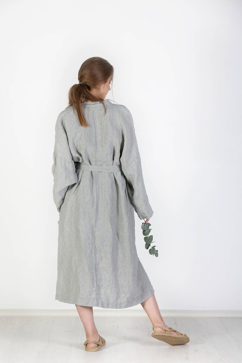 May include: A long, striped linen robe with a sash tie. The robe is a light gray color with thin, dark gray stripes. The robe is worn by a person standing with their back to the camera.