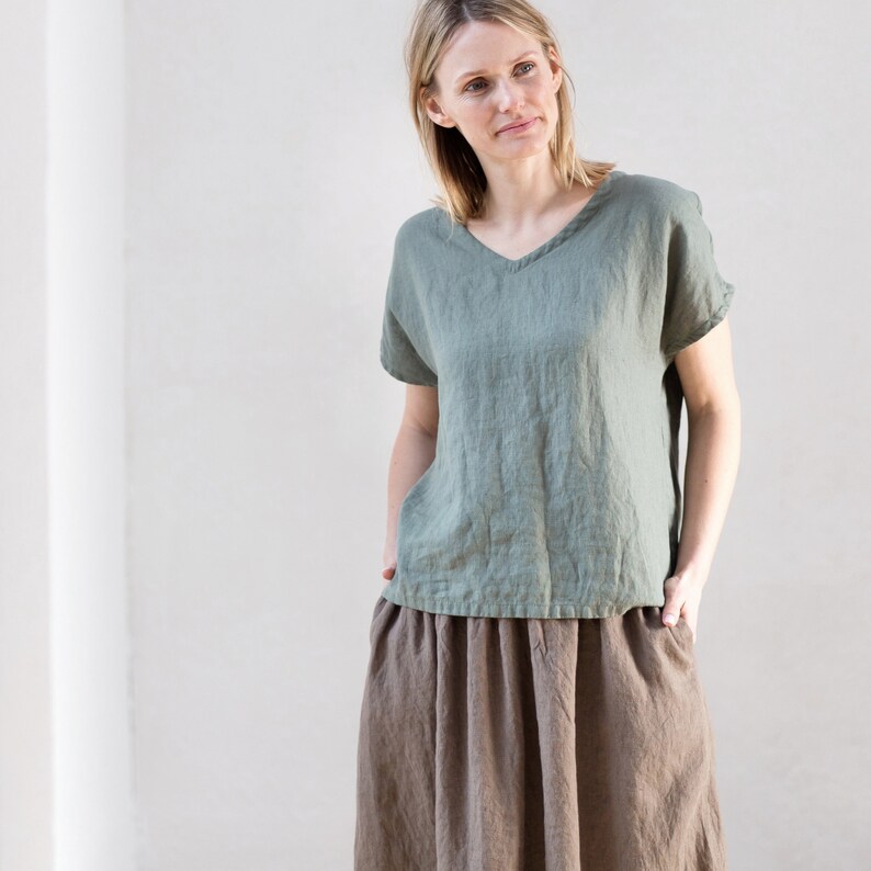 May include: A woman wearing a sage green linen top with a v-neck and a brown linen skirt.