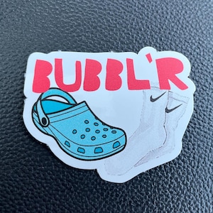May include: A white sticker with the text "BUBBLR'" in red letters. The sticker features a blue and white cartoon illustration of a clog and a pair of white socks with black Nike swooshes.