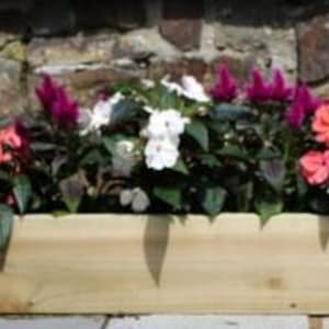 May include: A wooden rectangular planter box filled with colourful flowers. The flowers include shades of pink, white, and purple, with green foliage. The planter box is made of light-coloured wood and sits in front of a stone wall.