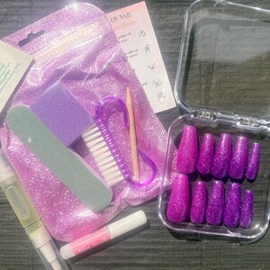 May include: A set of ten glittery purple press-on nails in a clear plastic case, accompanied by a purple glitter pouch, a nail file, a purple buffer block, a small nail brush, a wooden cuticle stick, a tube of nail glue, and a cuticle oil pen.