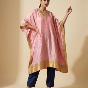 May include: A light pink kaftan featuring a V-neckline and wide sleeves, adorned with intricate gold metallic embroidery along the edges and hem. The garment is paired with solid navy blue trousers and light-colored pointed-toe heels.