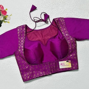 May include: A vibrant magenta blouse with short sleeves and a scoop neckline. The blouse features a gold patterned trim and a matching tie at the back. A small label is visible on the front. A bouquet of pink and white flowers is in the background.