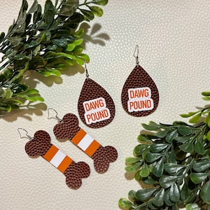 May include: A pair of brown and white football-themed earrings. One pair is shaped like a football and says "DAWG POUND" in white letters. The other pair is shaped like a bone and has white stripes.